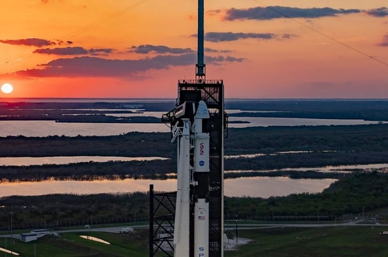 A SpaceX Falcon 9 rocket and spacecraft are pictured at a launch pad against a colorful sunset.